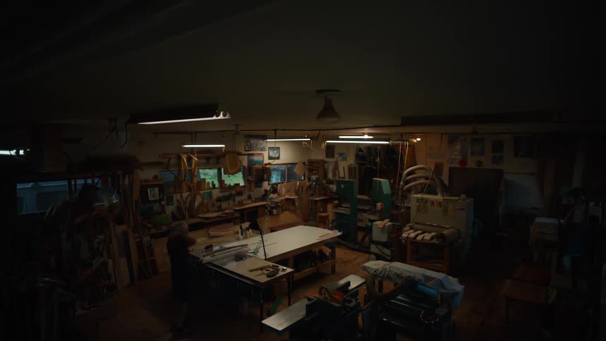 An elderly male carpenter walks to a table in a workshop