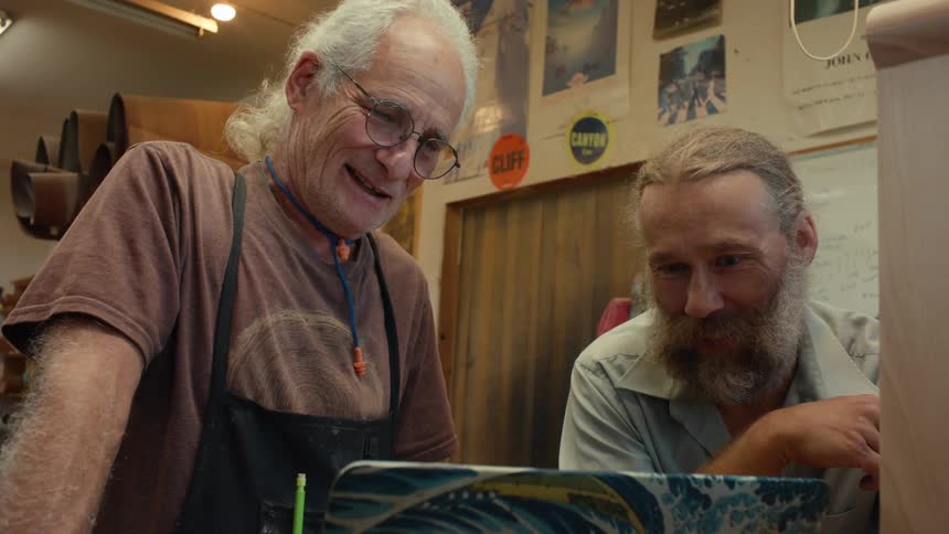 Two men in a workshop setting one wearing glasses and an apron smiling and engaged in conversation while looking at colorful material or artwork