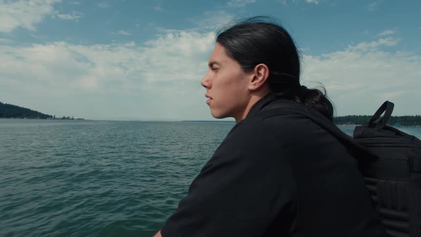 A man with long hair a black shirt and a backpack sits on a boat gazing thoughtfully out at the vast sea before him