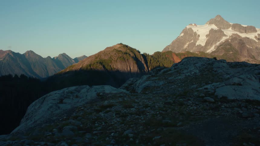 A rocky mountain peak casts a long shadow over a valley filled with rugged terrain and forested hills with a majestic snowcapped mountain visible in the distance