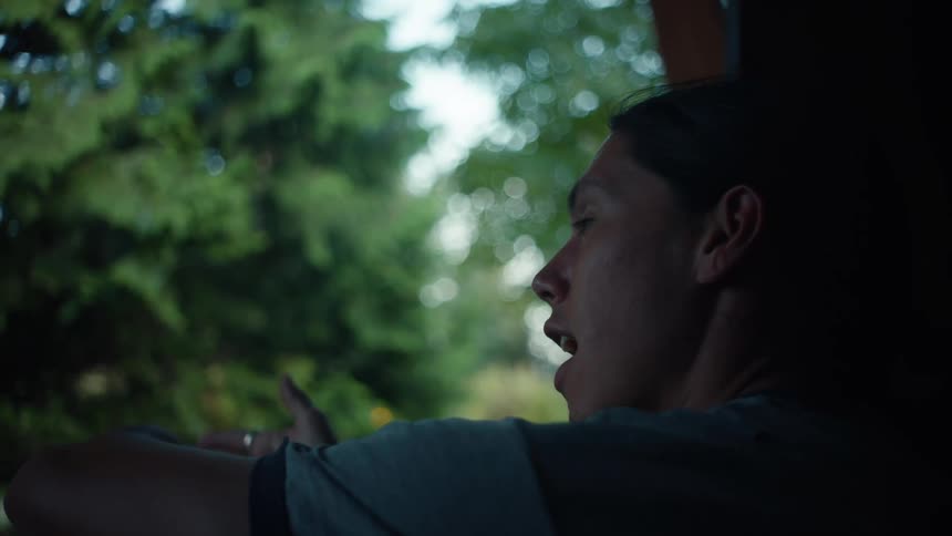 A person seen from the side looks out a window at a blurred view of trees while talking to someone