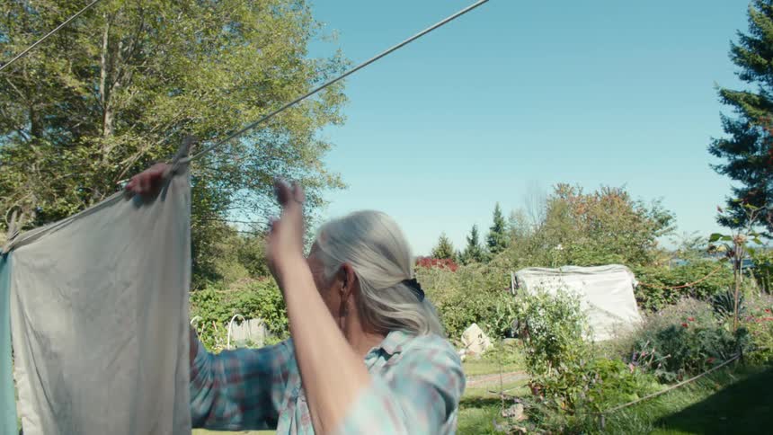 An older woman is hanging clothes on a clothesline in a sunny backyard garden