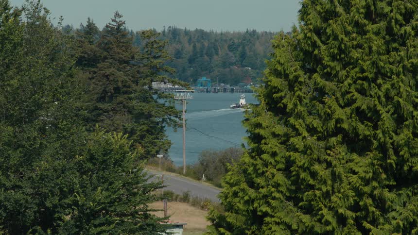A tugboat sails across a calm body of water and a small town are visible in the distance framed by lush green trees