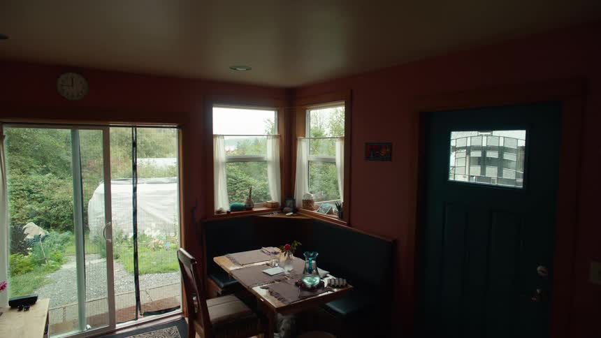 A room with a dining table in a corner with windows and on the left side there is a glass door that leads to a backyard and on the right side there is a blue door
