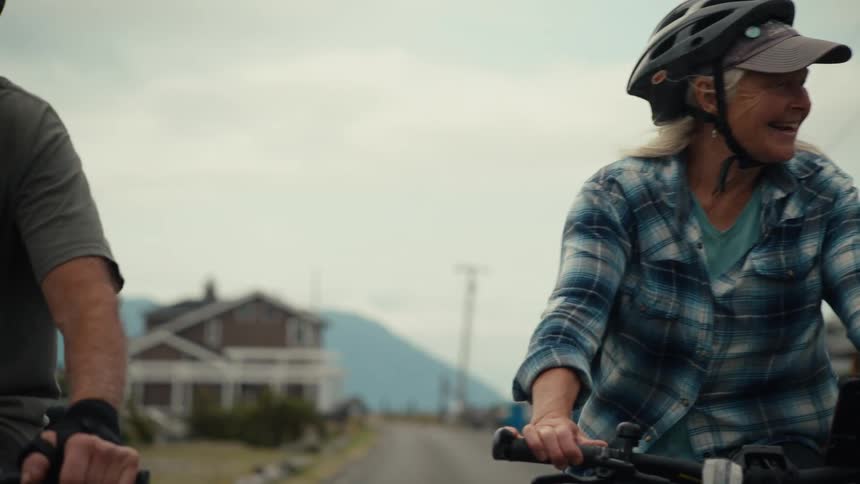 A woman and a man both wearing helmets are cycling down a road in front of houses