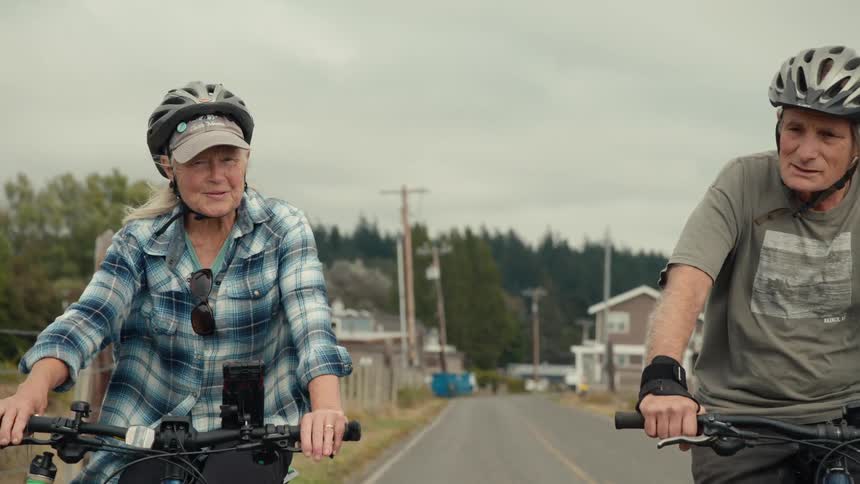 An elderly couple ride bikes together next to a lake and flatland
