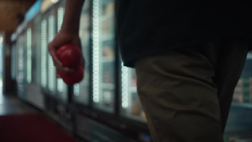 The man navigates the dimly lit aisle his hand tightly gripping a red tomato as he makes his way through the grocery store