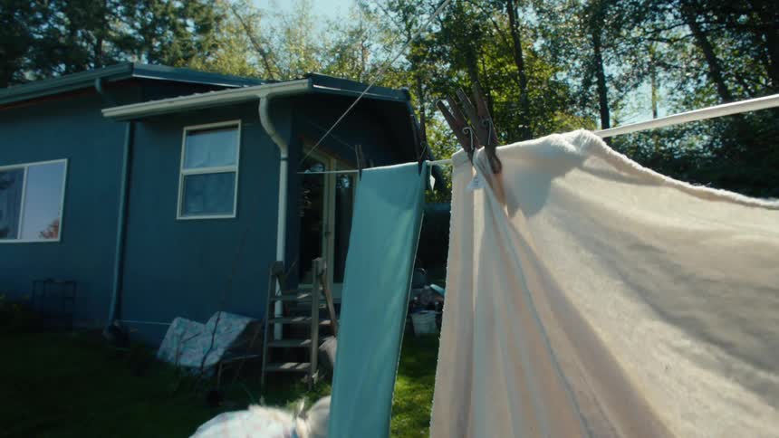 An elderly woman is hanging clothes on a clothesline outside her house surrounded by trees on a sunny day