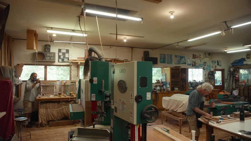 Two elderly men in a workshop are working with carpentry handling wooden objects and one of them is wearing a protective mask