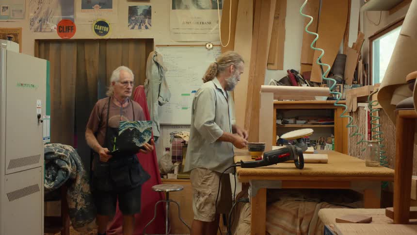 A carpenter is working on chair legs on his workbench when another carpenter enters the workshop carrying a laptop which he places on the workbench after moving the tools aside