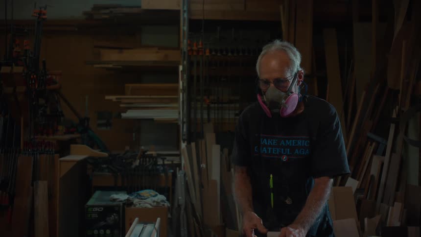 An older man wearing safety glasses and a respirator working in a woodworking shop filled with tools clamps and stacks of wooden planks