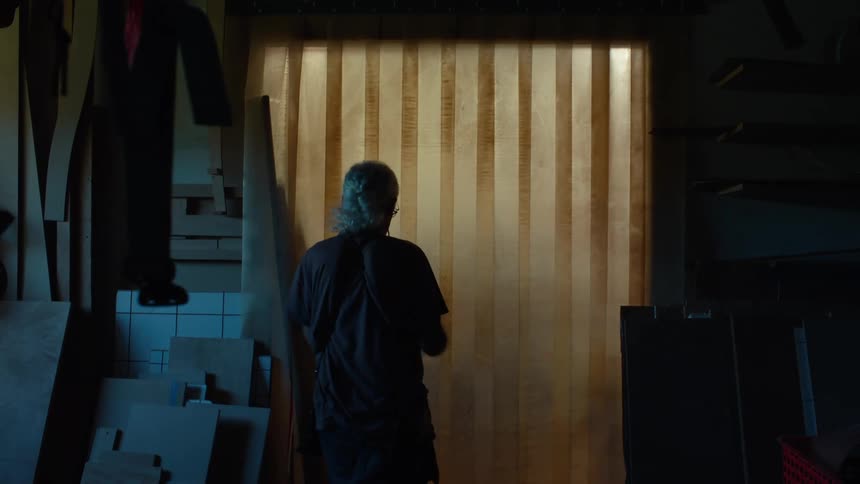 A person walks through vertical strips into a dimly lit workshop with wooden boards and materials stacked around