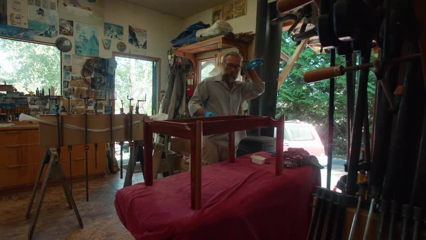 A man in a workshop carefully working on a piece of wooden furniture surrounded by tools clamps and posters with sunlight streaming through the windows