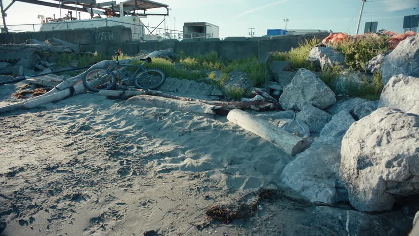 A rusted bicycle lies abandoned on a sandy shore littered with debris and large rocks with industrial structures looming in the background
