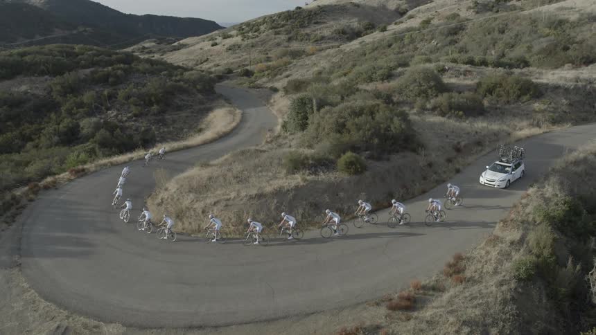 People on bicycles ride around a bending road followed by a car