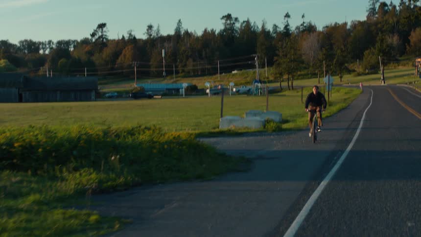 A cyclist riding on a rural road beside an open field with trees utility poles and buildings in the background under a clear sky