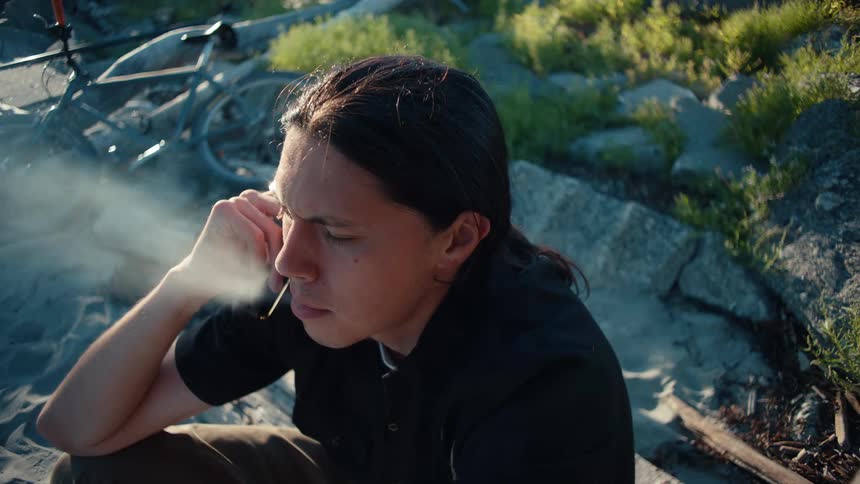 A person sits outdoors on a sandy surface smoking an ecigarette near rocks they hold a phone to their ear with a bicycle visible in the background