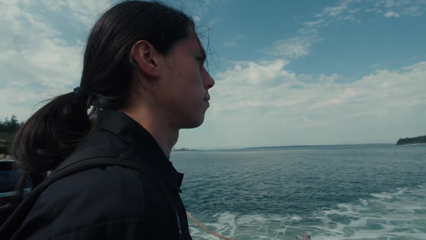 The face of a young man with ponytail looking at the sea as he stands on a ferry