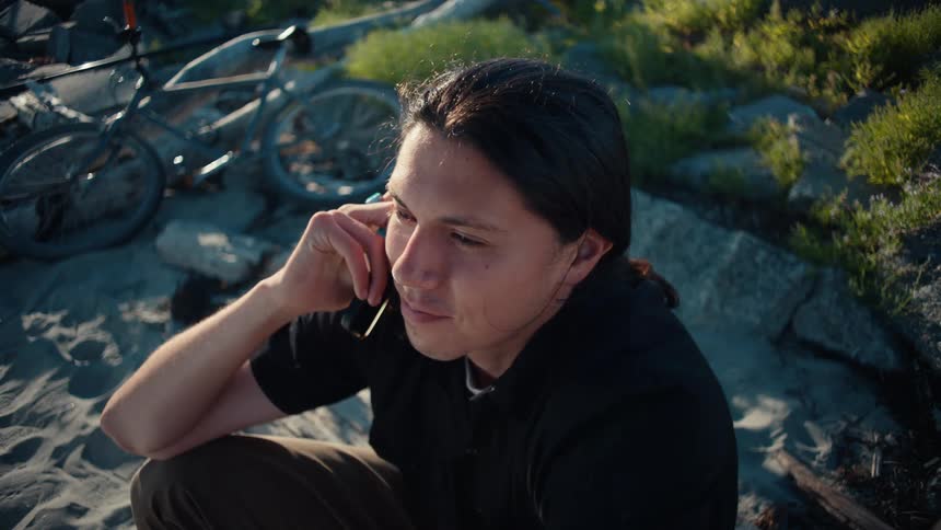 A man sitting outdoors on a sandy surface near rocks holding a phone to their ear with a bicycle visible in the background