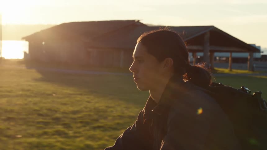 A man in a black jacket and backpack rides a bicycle along a rural road with a basketball court and playground visible in the background as the sun sets