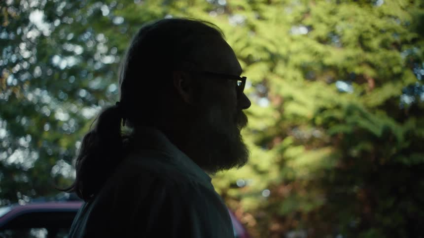 A bearded man with flowing hair silhouetted against the backdrop of a sunlit forest and a vibrant red car is deep in conversation