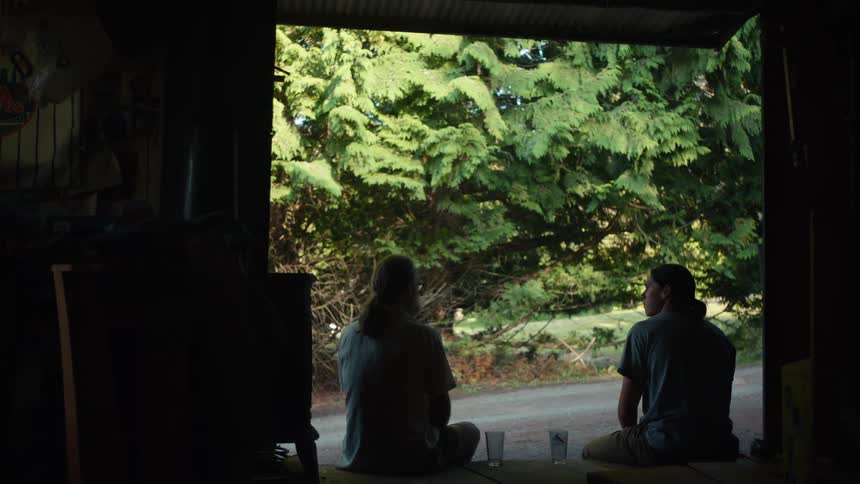 Two people sit on the floor of the open doorway of a workshop facing a sunlit area with dense folliage
