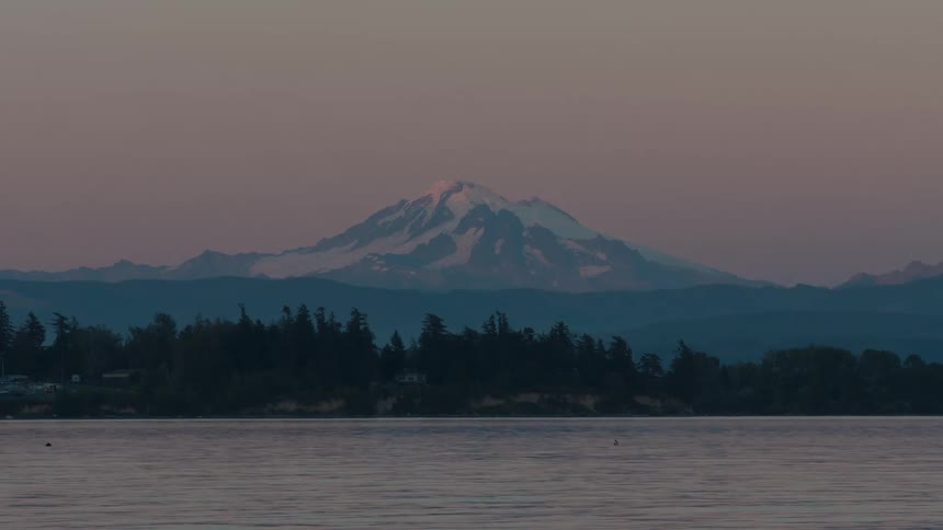 A snowcapped mountain stands tall against a soft pink sky reflecting in the calm waters of a lake
