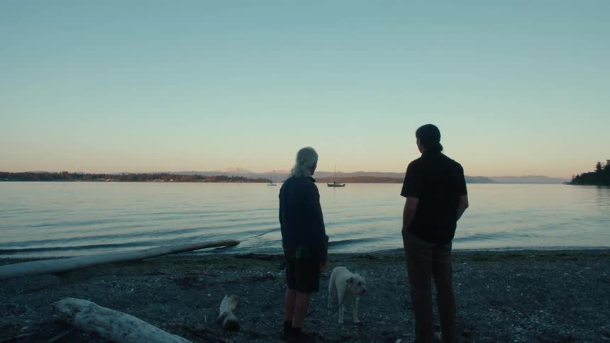 A man and a teenager talk on a beach as boats float in the water