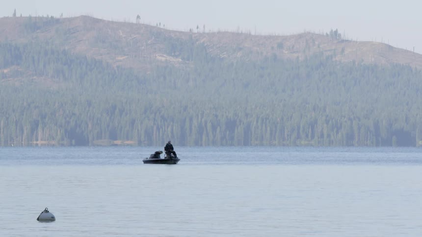 A man sitting on the front of a small boat, in the middle of a pond, fishing