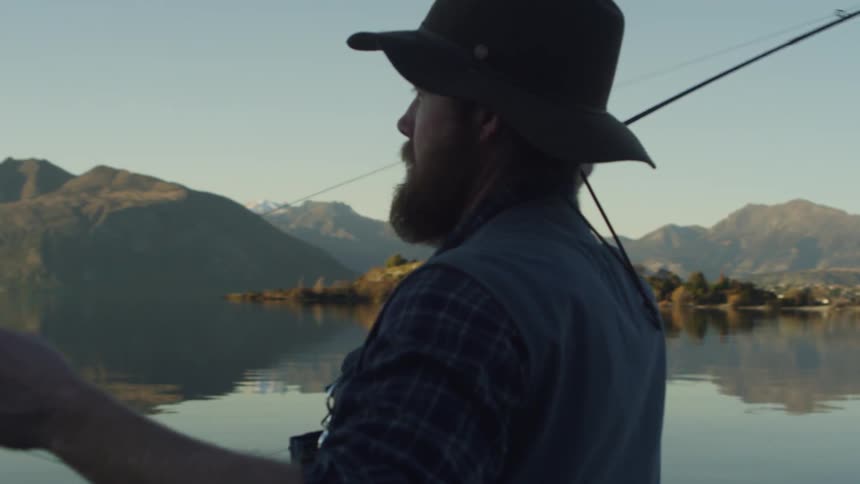 A man in fishing gear casts his fishing line into a calm lake surrounded by mountains