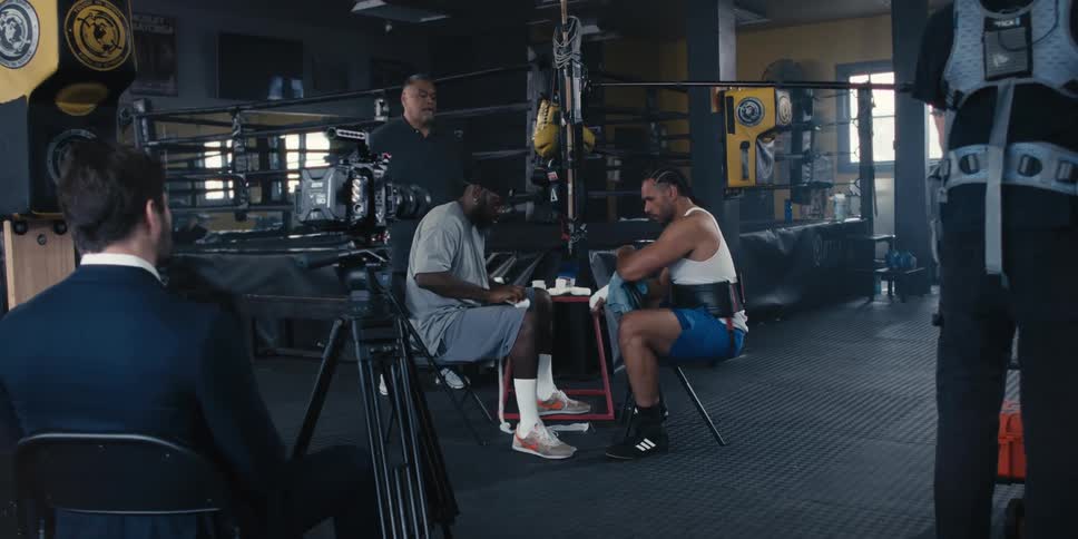 Boxer sitting with another man bandaging his hand and around a man in a blazer with a camera