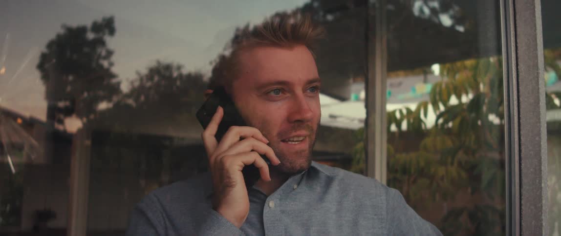 The face of a young blond man who is talking on his cell phone which is at his ear and he is behind a glass window