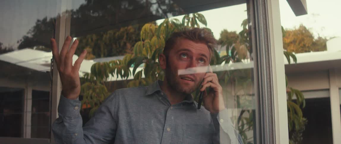 A young man in a dress shirt is talking on the phone that is held to his ear and he is pointing to the sky while standing inside a room seen through a glass window