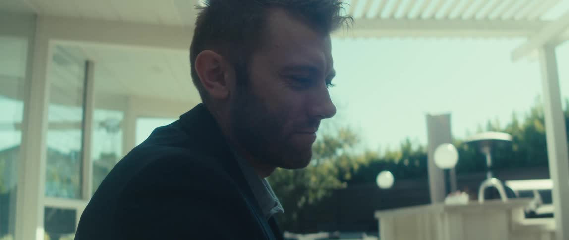 A person in a dark suit jacket and lightcolored shirt sits outdoors in the backyard at home