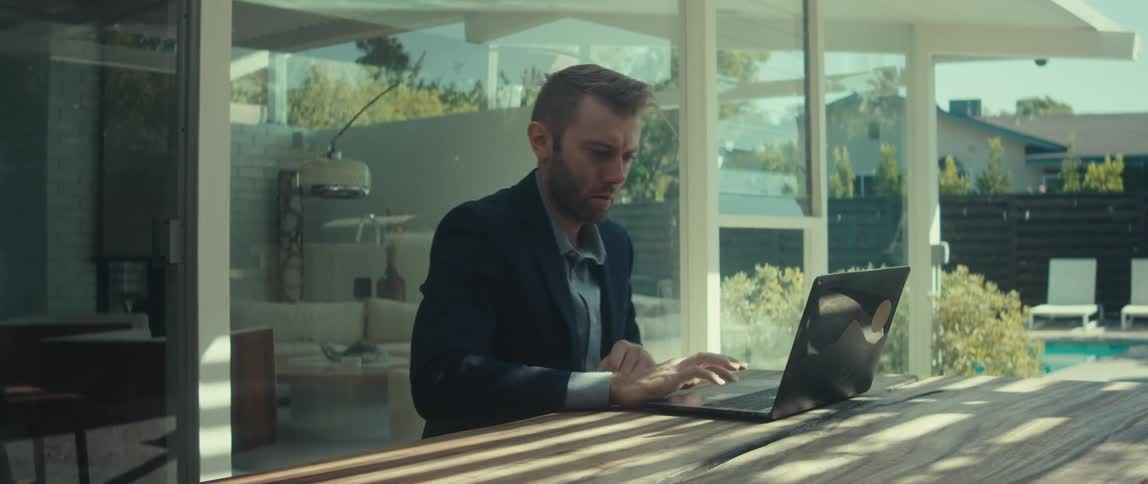 A man in a dark suit jacket and lightcolored shirt sits outdoors working on his laptop