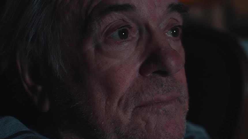 The face of an elderly man who is pointing a remote control at a screen that is shining in his face
