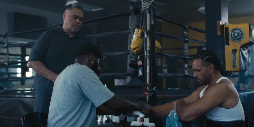 In a boxing gym setting a person is carefully wrapping a boxers hands in white bandages while another man watches