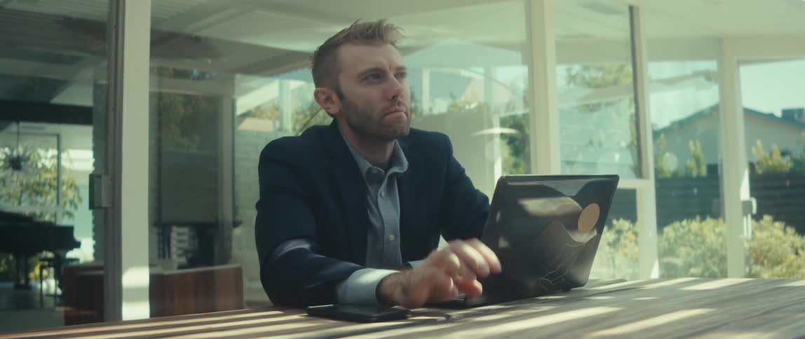 Blonde man sitting at a wooden table outside interacting with his laptop