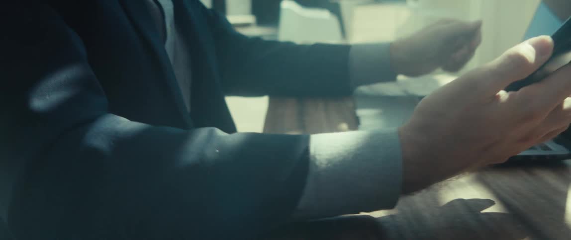 A man in a dark suit jacket and lightcolored shirt sits outdoors working on his laptop and checking his phone
