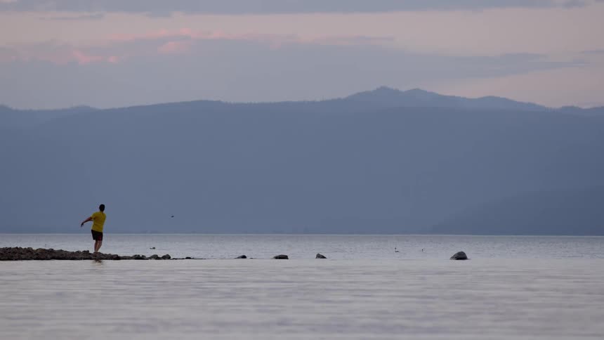 A man standing on some rocks at the edge of water and throwing rocks in