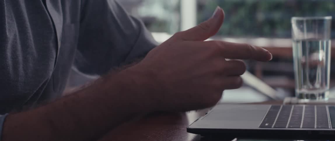 A man in a lightcolored shirt sits outdoors having a meeting on his laptop