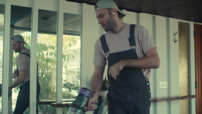 A man in a cap is cleaning a black floor with a vacuum and he is moving in dance tones