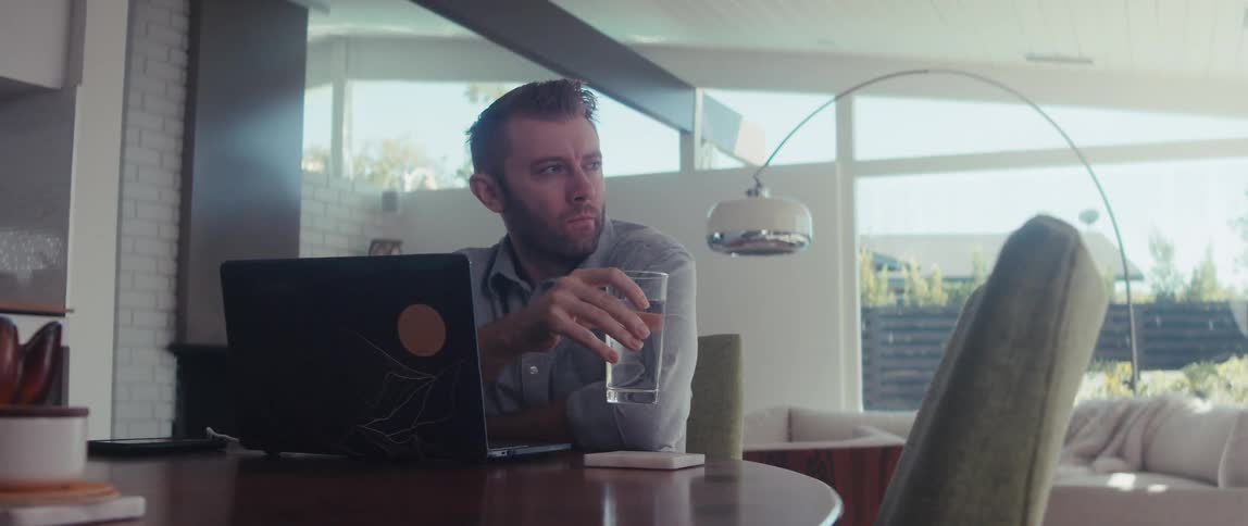 A man is sitting at a living room table with a pensive look drinking from a glass of water then puts the glass down and turns to his laptop