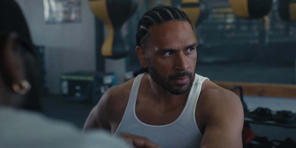 The face of a man with braids in his hair and wearing a black tank top who is looking straight ahead where there is another man fiddling with something and then he looks to his left side