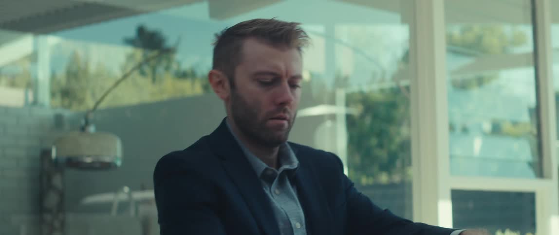 A man in a dark suit jacket and lightcolored shirt sits outdoors working on his laptop