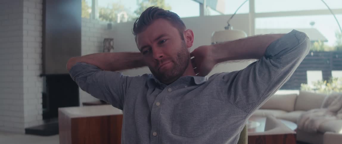 A man in a shirt sits stretching on a welllit living room in a modern home
