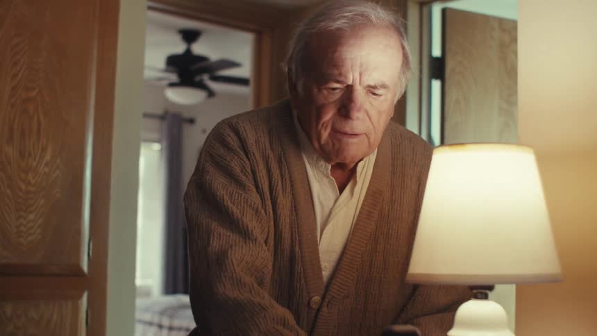 An elderly man sitting on a bed stands up to answer the phone at home