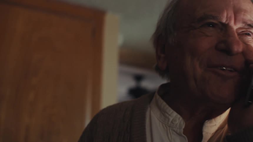 An elderly man picks up a call on a cordless phone and smiles and laughs as he talks