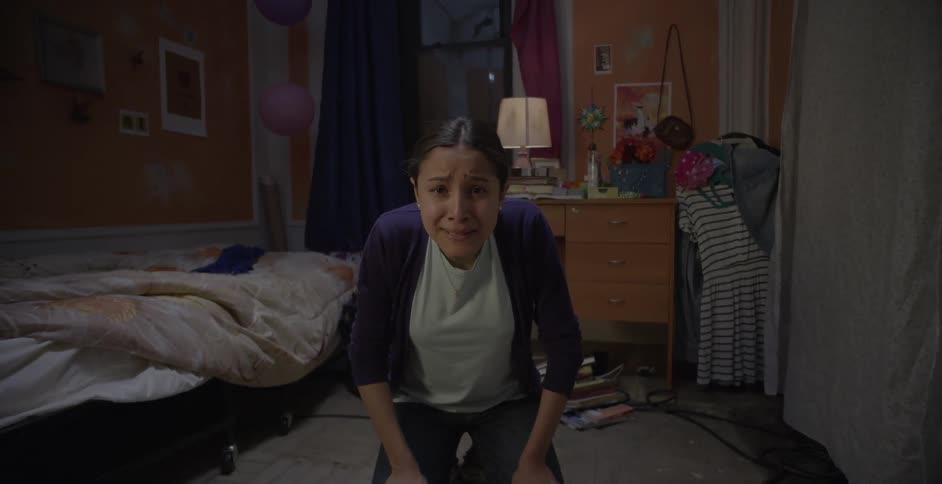 A woman kneels on the ground in a bedroom and is crying