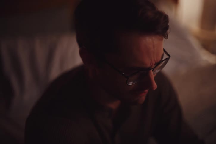 A man clad in sunglasses and a black sweater sits on the bed